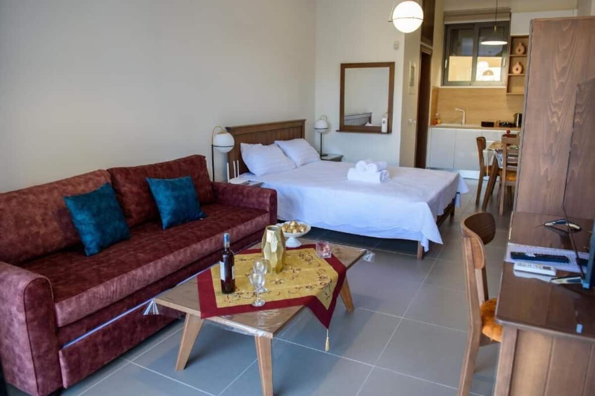 A hotel room with a red sofa, a bed with white linens, a small dining table with a bottle and glasses, and a kitchenette in the background.