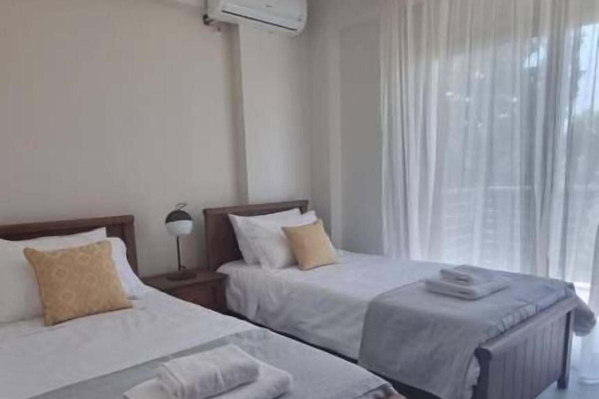 A hotel room with two beds, a ceiling fan, an air conditioning unit, and a large window with sheer curtains.