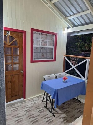 A small room with a wooden door, a window with a red frame, a table with a blue tablecloth, and a chair. - THE COZY WOODEN NEST IN LABORIE (LABORIE)