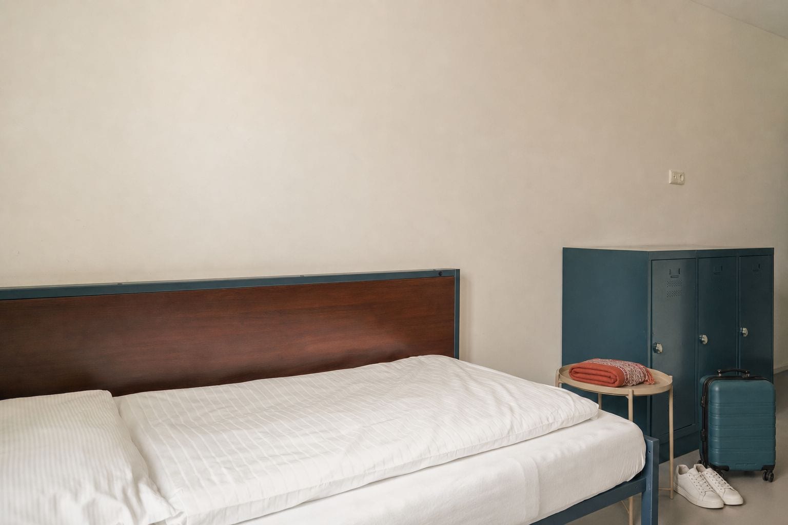 Un lit simple avec une tête de lit en bois, une petite table ronde recouverte d’une nappe à motifs rouges et blancs, une valise bleue et une paire de chaussures blanches.