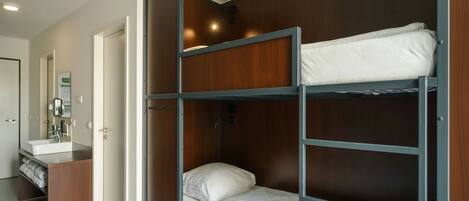 Una cama individual con estructura de madera y ropa de cama blanca, un baño con lavabo y perchero, y una puerta que da a otra habitación.