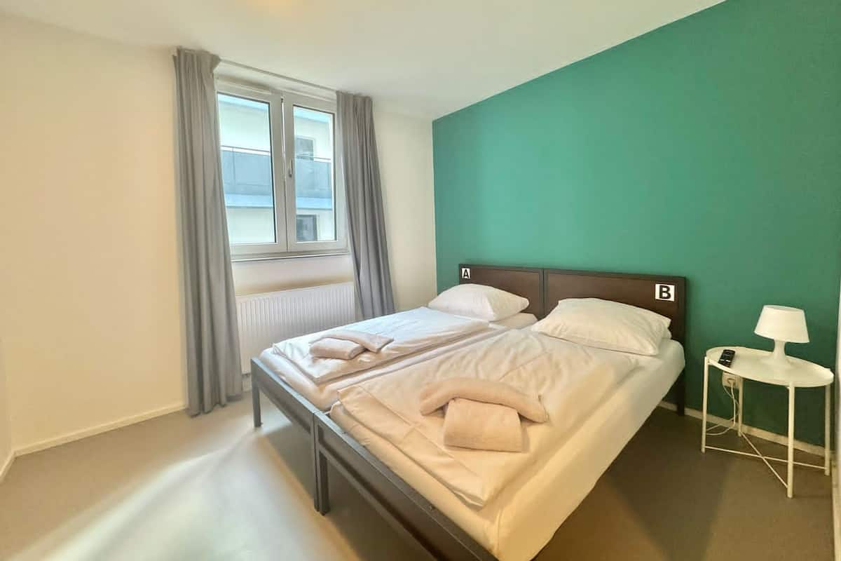 A single-bed room with a wooden headboard, a window with curtains, a bedside table with a lamp, and a green accent wall.