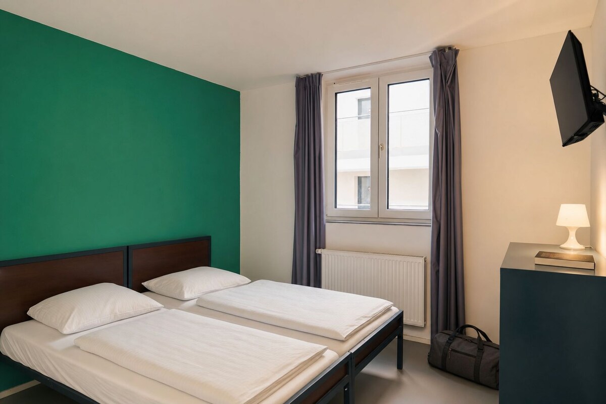 Une chambre d’hôtel avec deux lits, une fenêtre avec des rideaux, un radiateur, un bureau avec une lampe et un téléviseur fixé au mur.