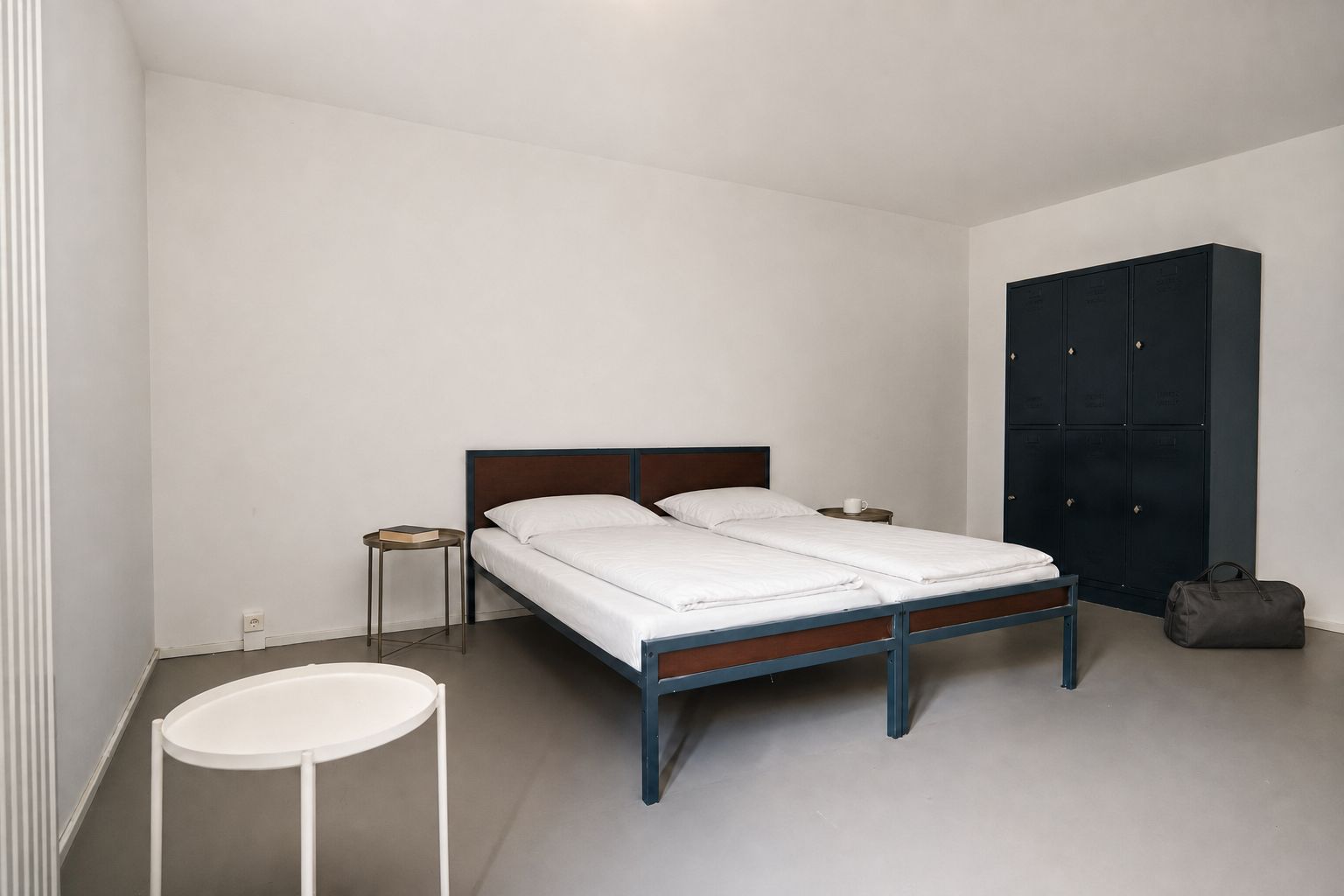 Una cama individual con estructura de madera y ropa de cama blanca, una mesita de noche con un libro, un taburete redondo blanco y un armario de color oscuro.