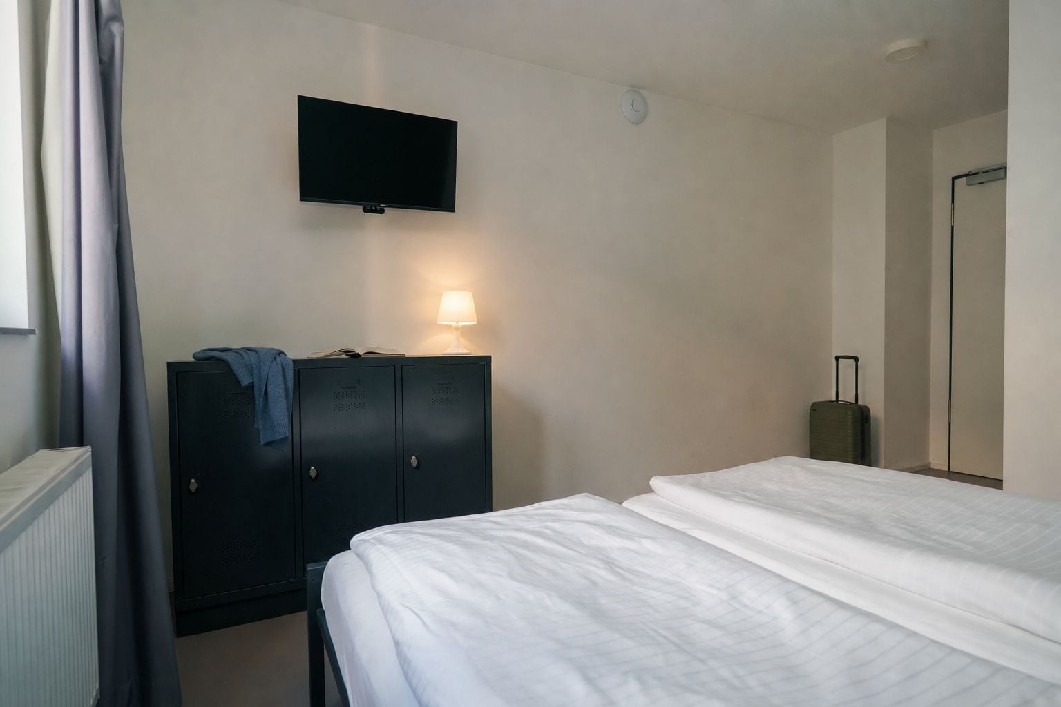 Una habitación de hotel con una cama, un televisor montado en la pared, un armario y una maleta.