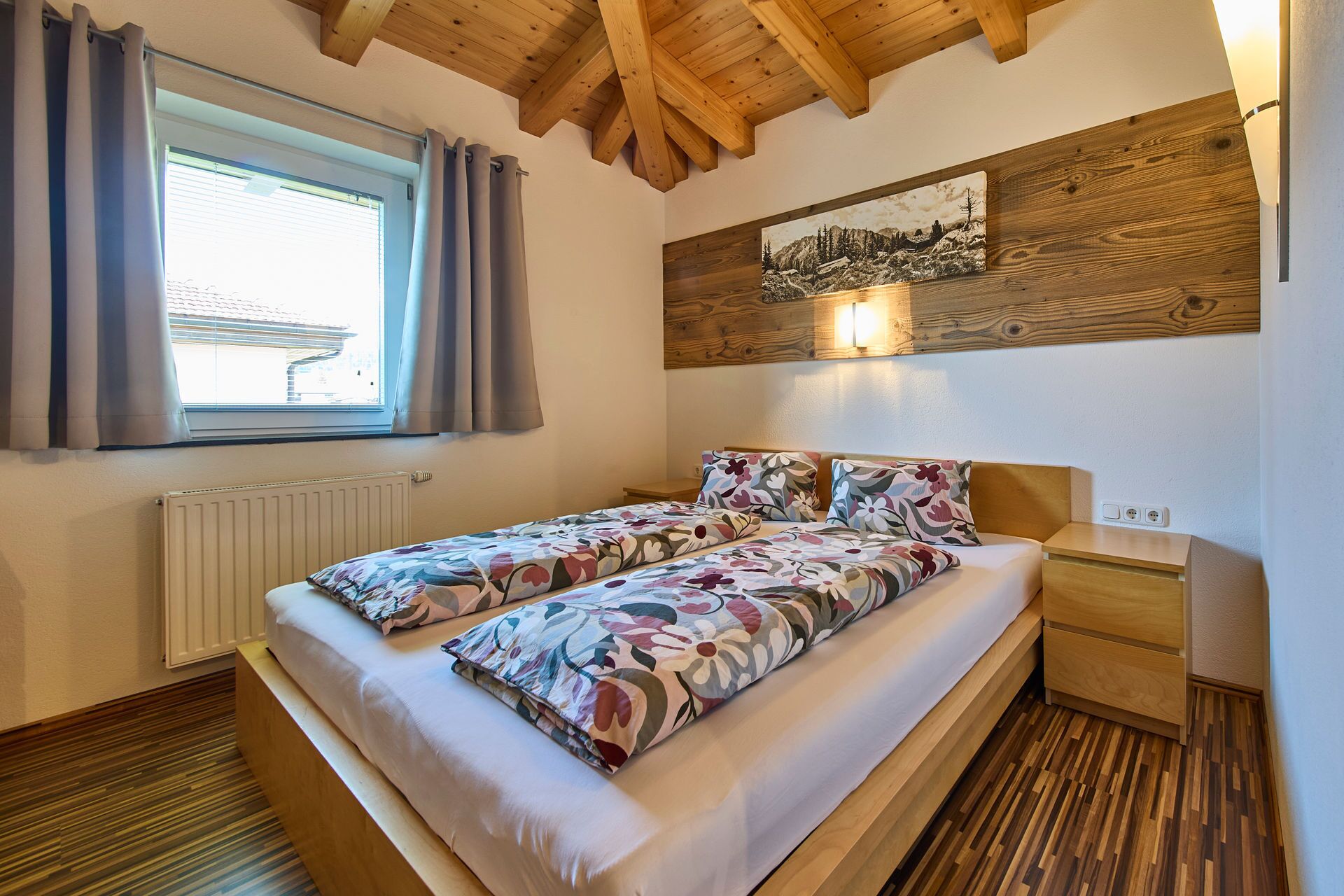 A bedroom with a wooden ceiling, a bed with floral bedding, a nightstand, a wall-mounted light, and a window with curtains.