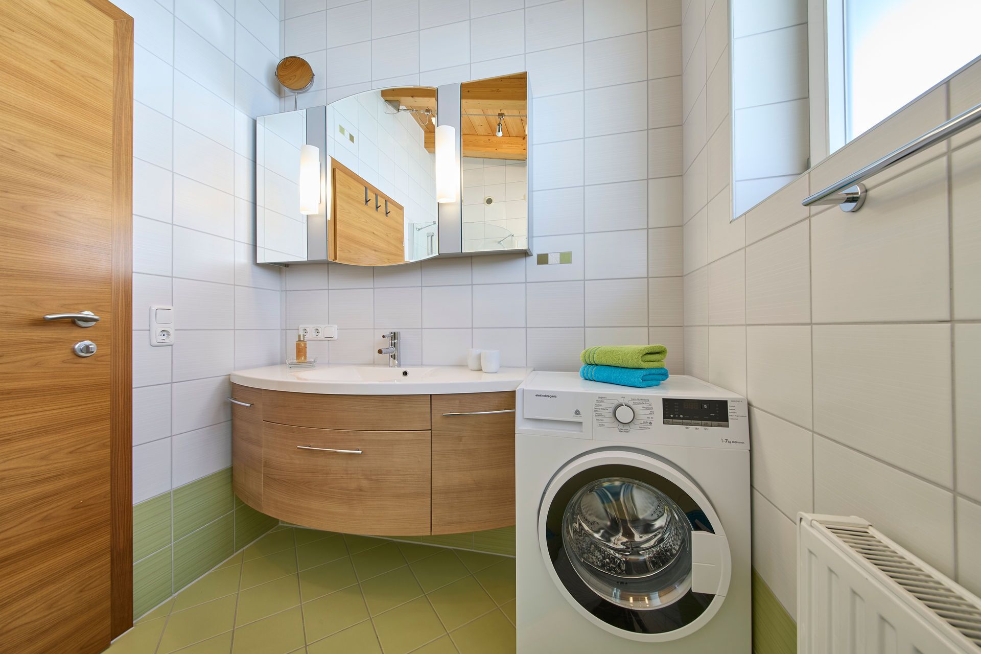 Ein kleines Badezimmer mit Holztür, einem weißen Waschbecken mit Spiegel darüber, einer integrierten Waschmaschine und einem Heizkörper.