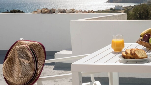 A white table with a glass of orange juice, a plate of bread, and a straw hat on a balcony overlooking the sea. -  Deluxe apartment with panoramic sea view located on the hill of Provatas (Milos)