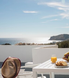 Une table blanche avec un verre de jus d’orange, une assiette de pain et un chapeau de paille sur un balcon donnant sur la mer.