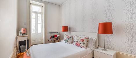 A bedroom with a bed, two bedside tables, a nightstand, a chair, a desk with a computer, and a radiator.