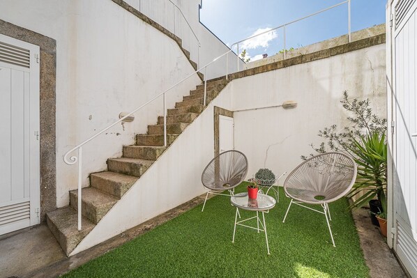 Una escalera que lleva a una puerta blanca, un pequeño espacio al aire libre con dos sillas y una mesa, y una planta en maceta.