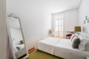 A hotel room with a large mirror, a bed with white bedding, a bedside table with a lamp, and a view of the outside through a window.