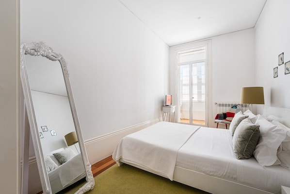 Une chambre d’hôtel avec un grand miroir, un lit avec une literie blanche, une table de chevet avec une lampe et une vue sur l’extérieur par une fenêtre.