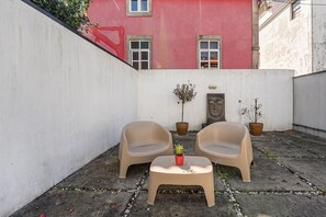 Uma pequena área externa com assentos, incluindo duas cadeiras de plástico bege e uma mesa pequena, cercada por plantas em vasos e uma parede de pedra.