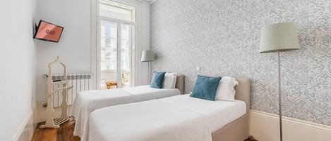A hotel room with two beds, a radiator, a desk, and a window.