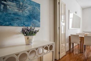 Une pièce moderne avec une console blanche, un pot de lavande, un tableau abstrait bleu, un coin repas avec des chaises blanches et un grand miroir.