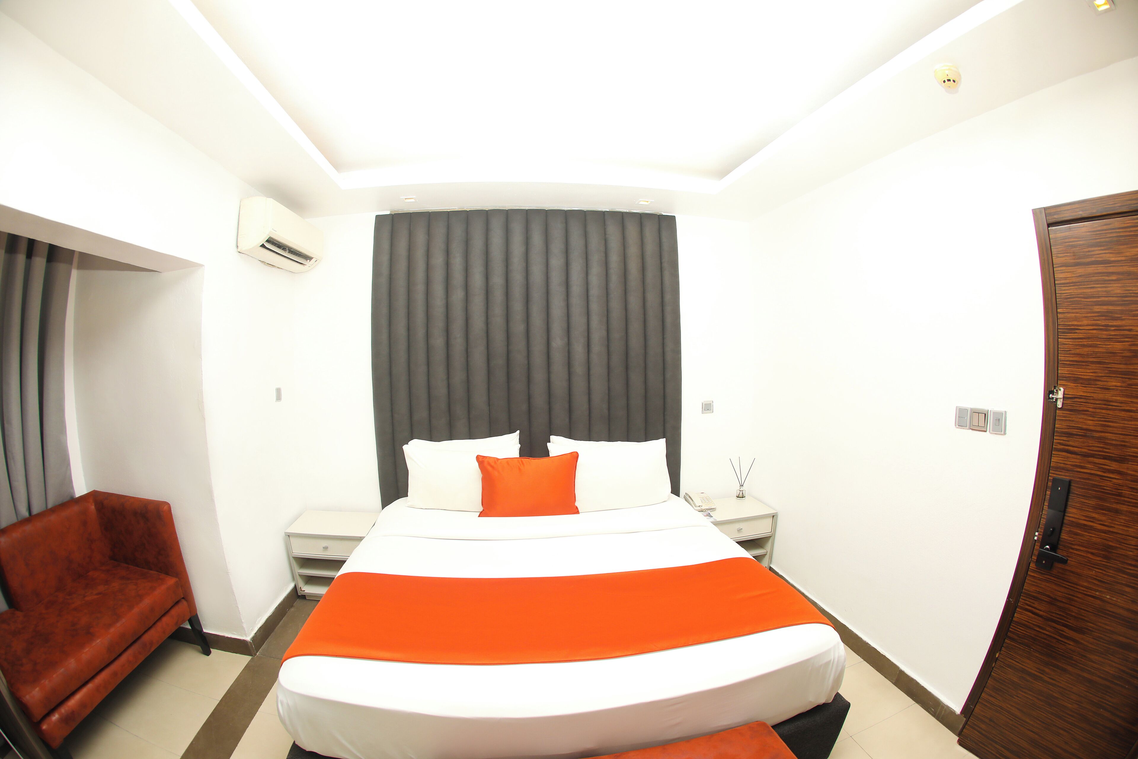 A hotel room with a large bed, a red armchair, a bedside table, and a wooden door.