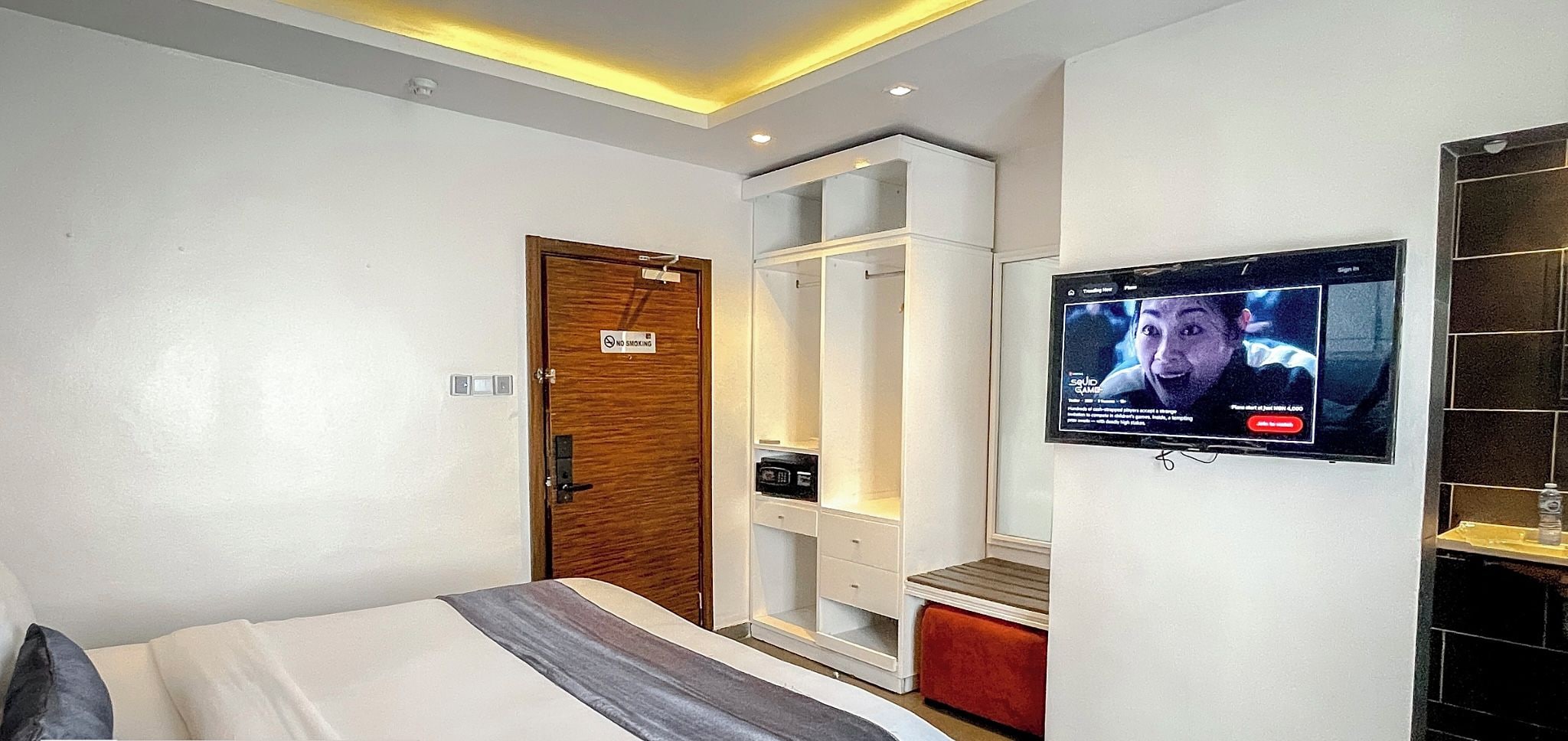 A hotel room with a bed, a TV mounted on the wall, a wardrobe, and a "No Smoking" sign on the door.