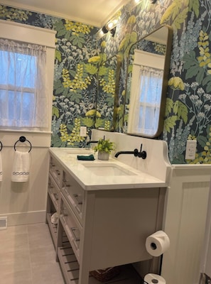Casa de banho com papel de parede floral, uma pia branca, um espelho e um porta-toalhas.
