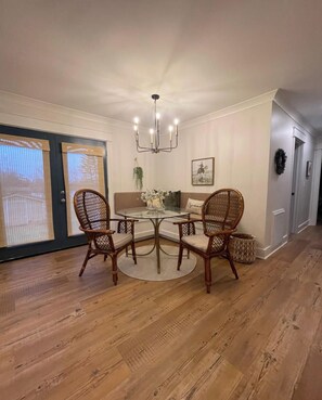 Un coin repas avec une table ronde, deux chaises en osier, un lustre et une vue sur l’extérieur grâce à de grandes fenêtres.
