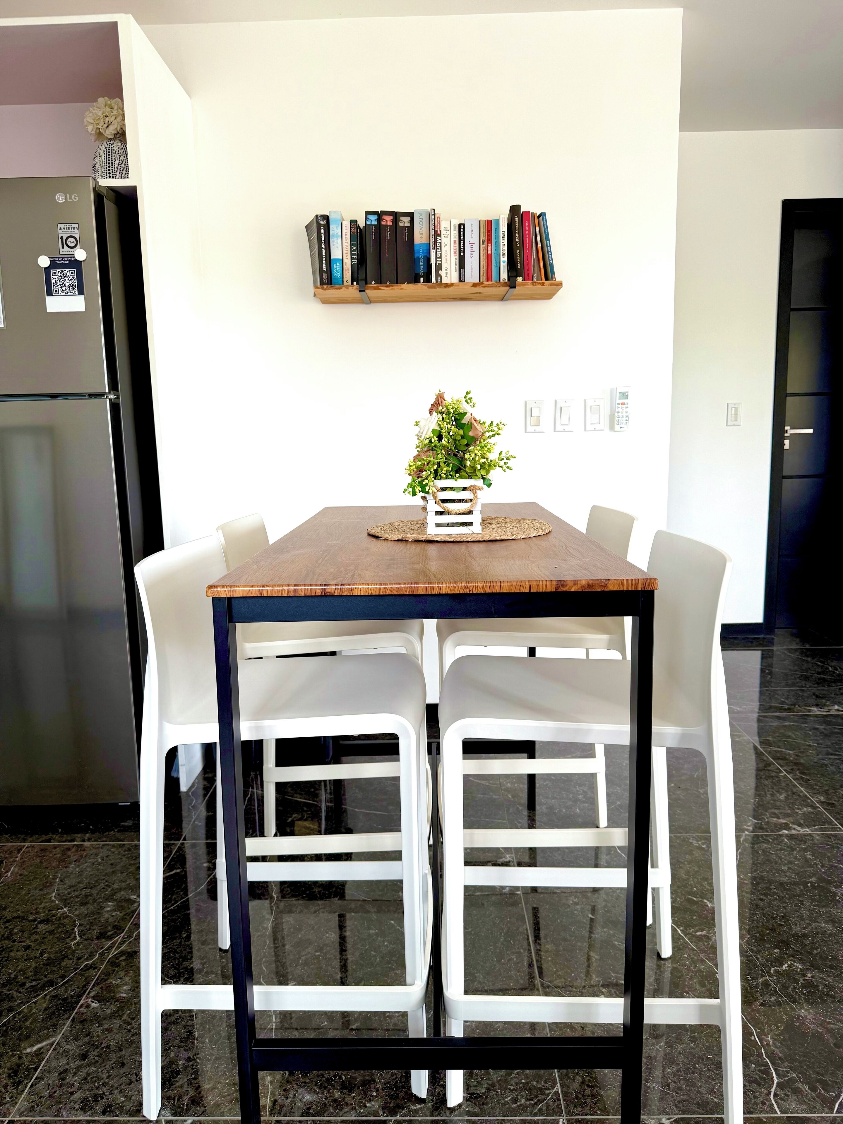 A modern kitchen with a dining table, white chairs, a wooden shelf with books, and a decorative plant.