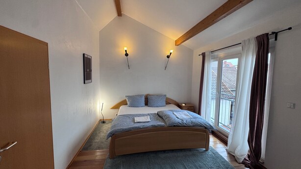 Een slaapkamer met een houten bed, een klein nachtkastje, gordijnen voor het raam en uitzicht op gebouwen.