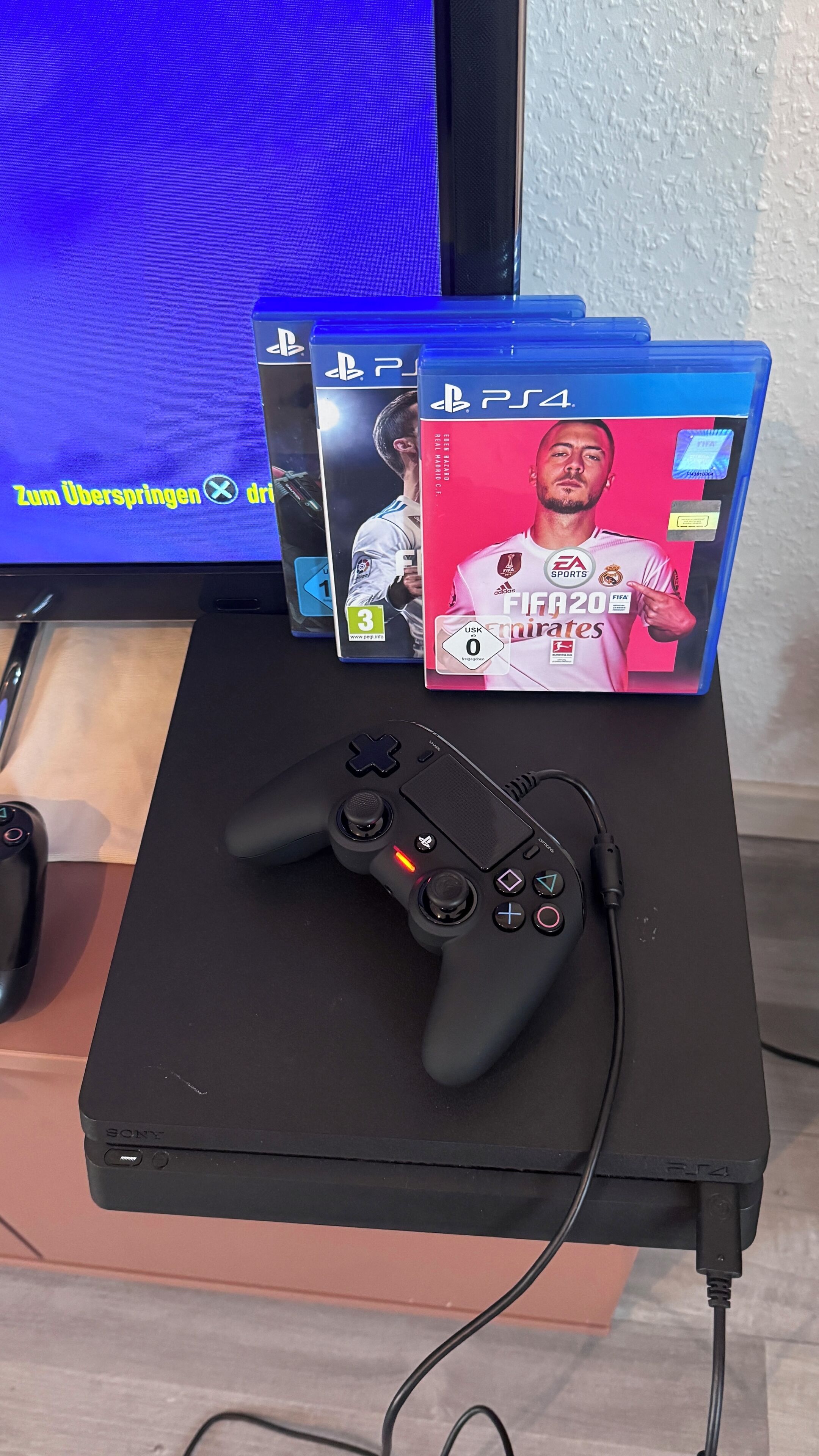 Een gaming-opstelling met een PlayStation 4 console, twee spelletjesdoosjes en een zwarte game controller op een zwarte standaard.