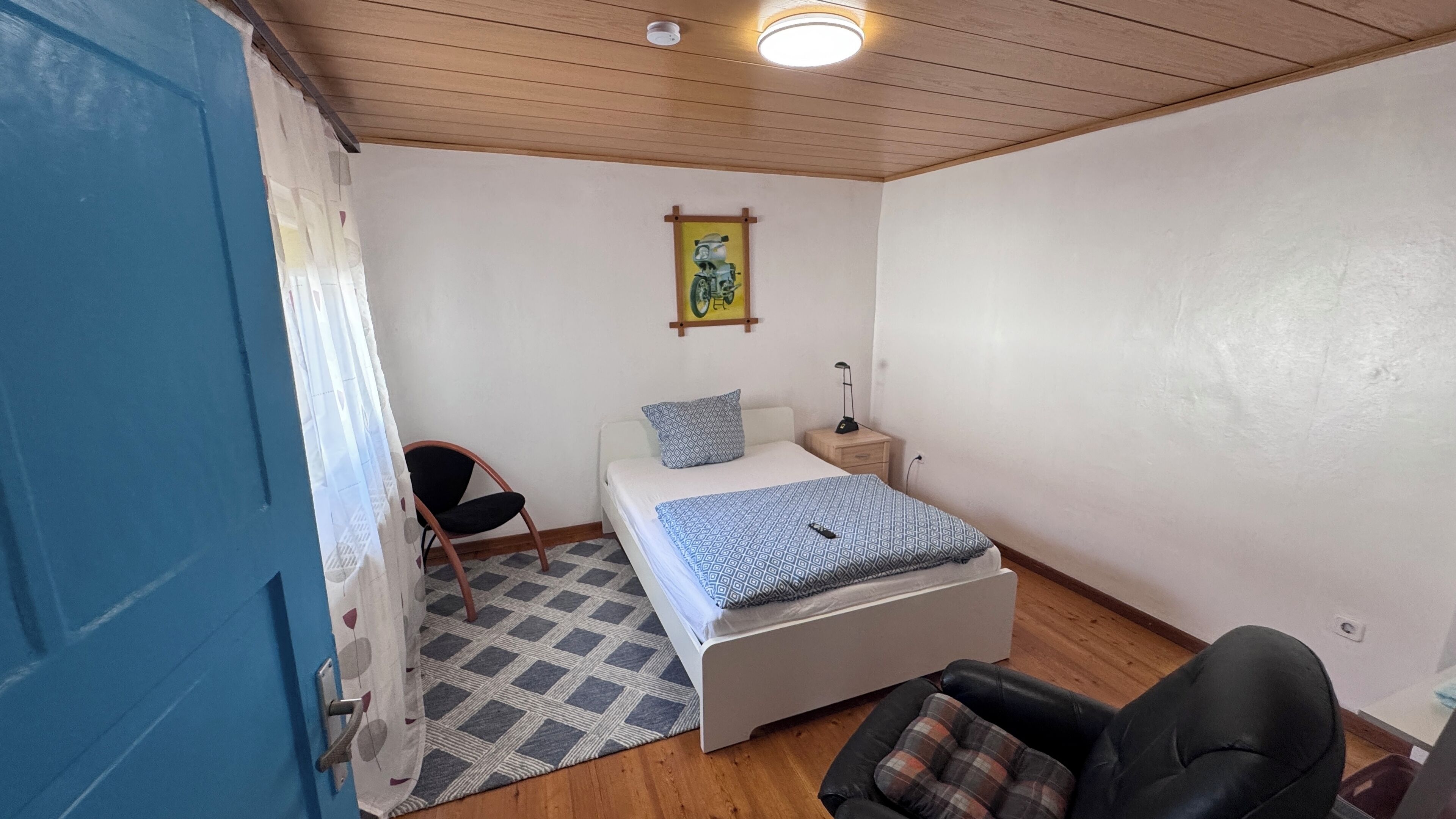A small, well-lit bedroom with a bed, a chair, a nightstand, and a painting on the wall.