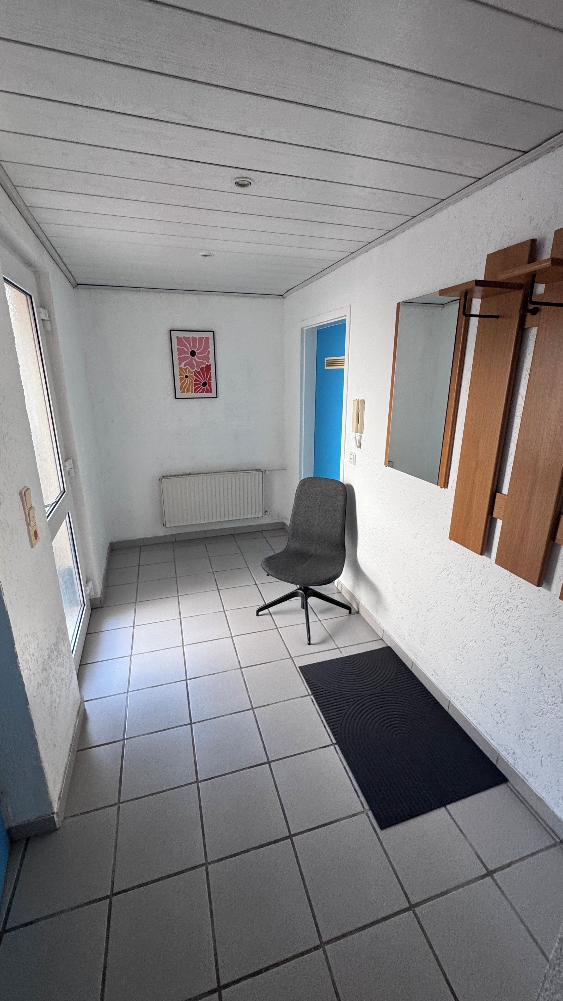 Ein Zimmer mit Fliesenboden, einem grauen Bürostuhl, einem schwarzen Teppich, einem Heizkörper, einer blauen Tür und einem gerahmten Bild an der Wand.