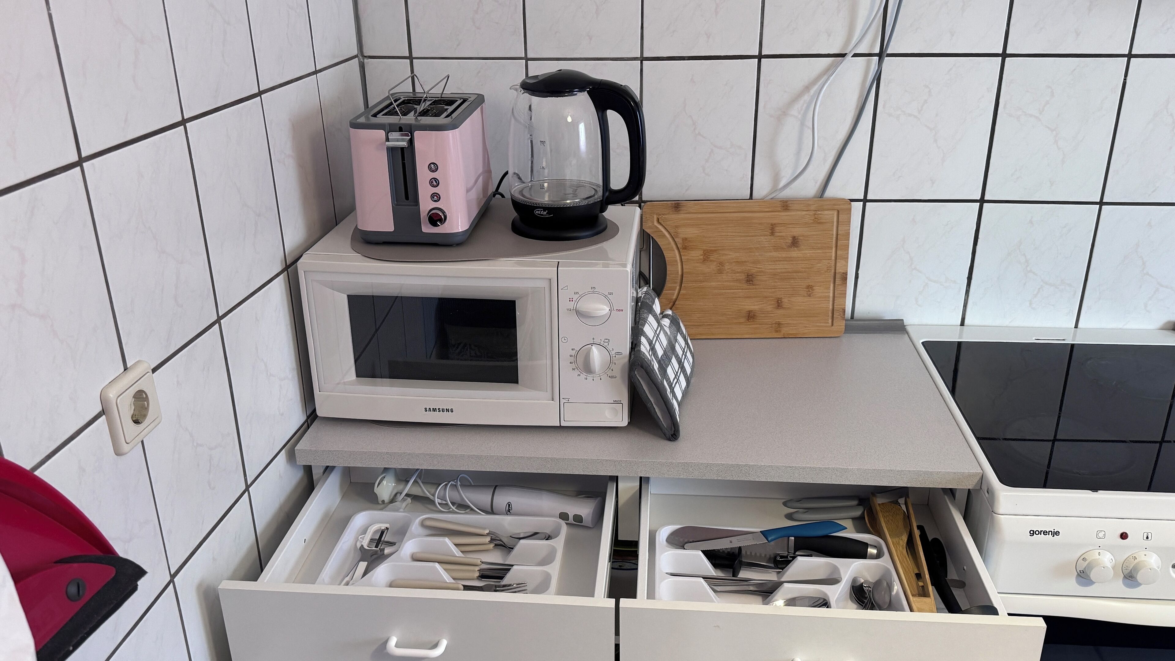 A kitchen with a microwave, toaster, and kettle on the countertop, a wooden cutting board, and a drawer with utensils.