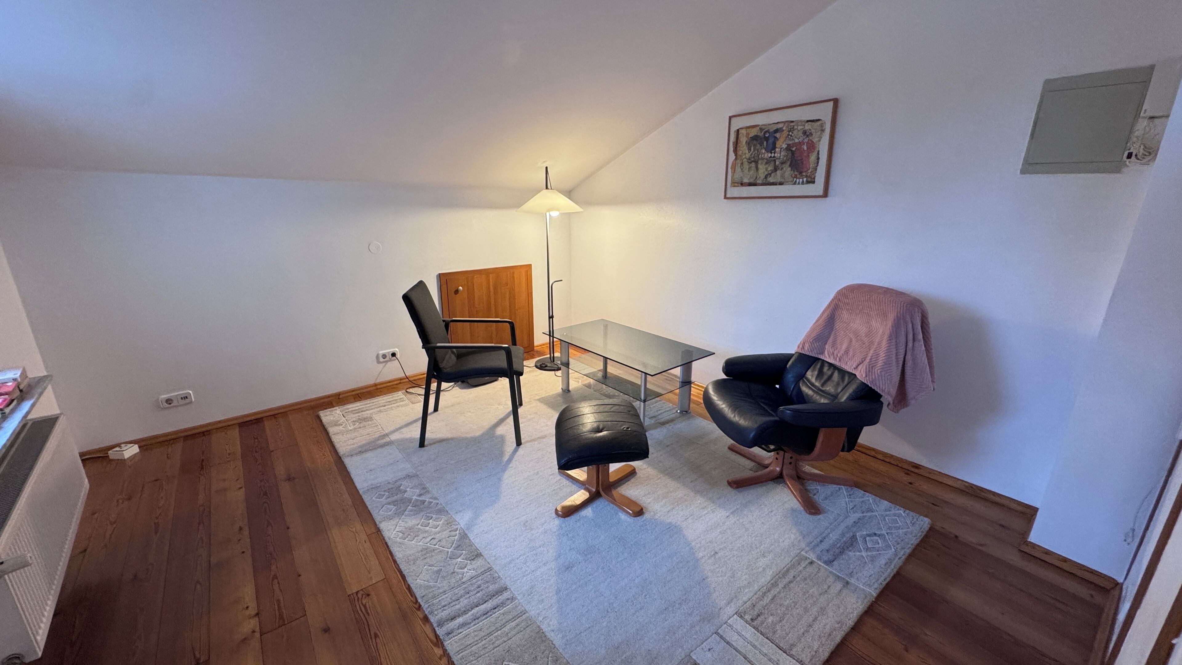 Ett litet, väl upplyst rum med ett trägolv, ett glasbord, två stolar, en bänk, en golvlampa och en tavla på väggen.