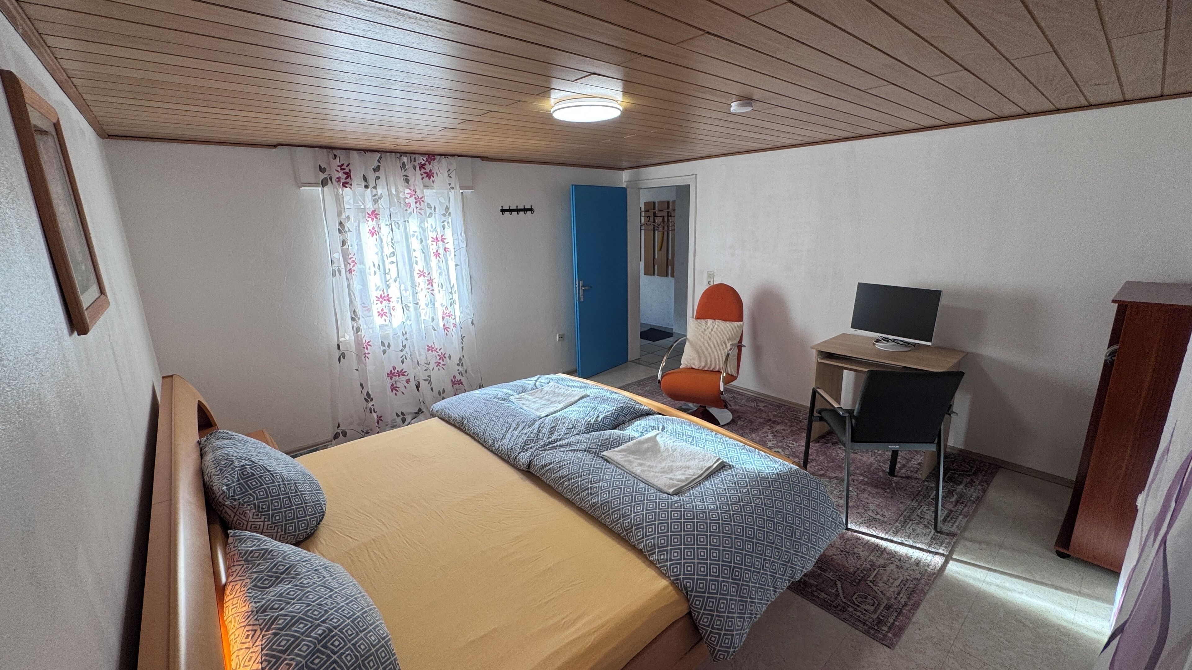 A bedroom with a bed, a desk with a computer, a chair, and a wardrobe.