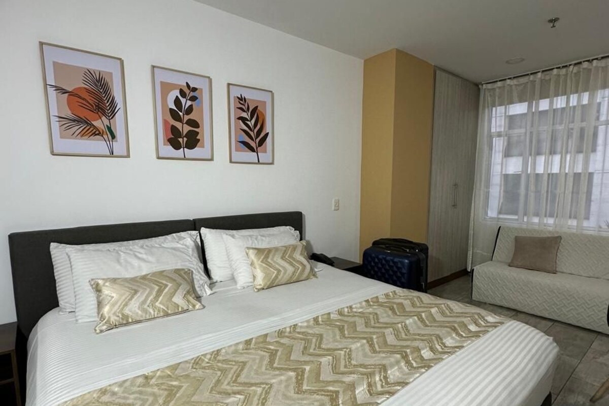 A hotel room with a large bed, a sofa, a suitcase, and three framed wall art pieces.