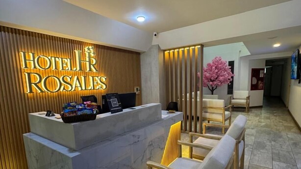 A hotel reception area with a marble reception desk, a neon sign, and a seating area with wooden chairs. - Hotel Rosales Suites (Pereira)