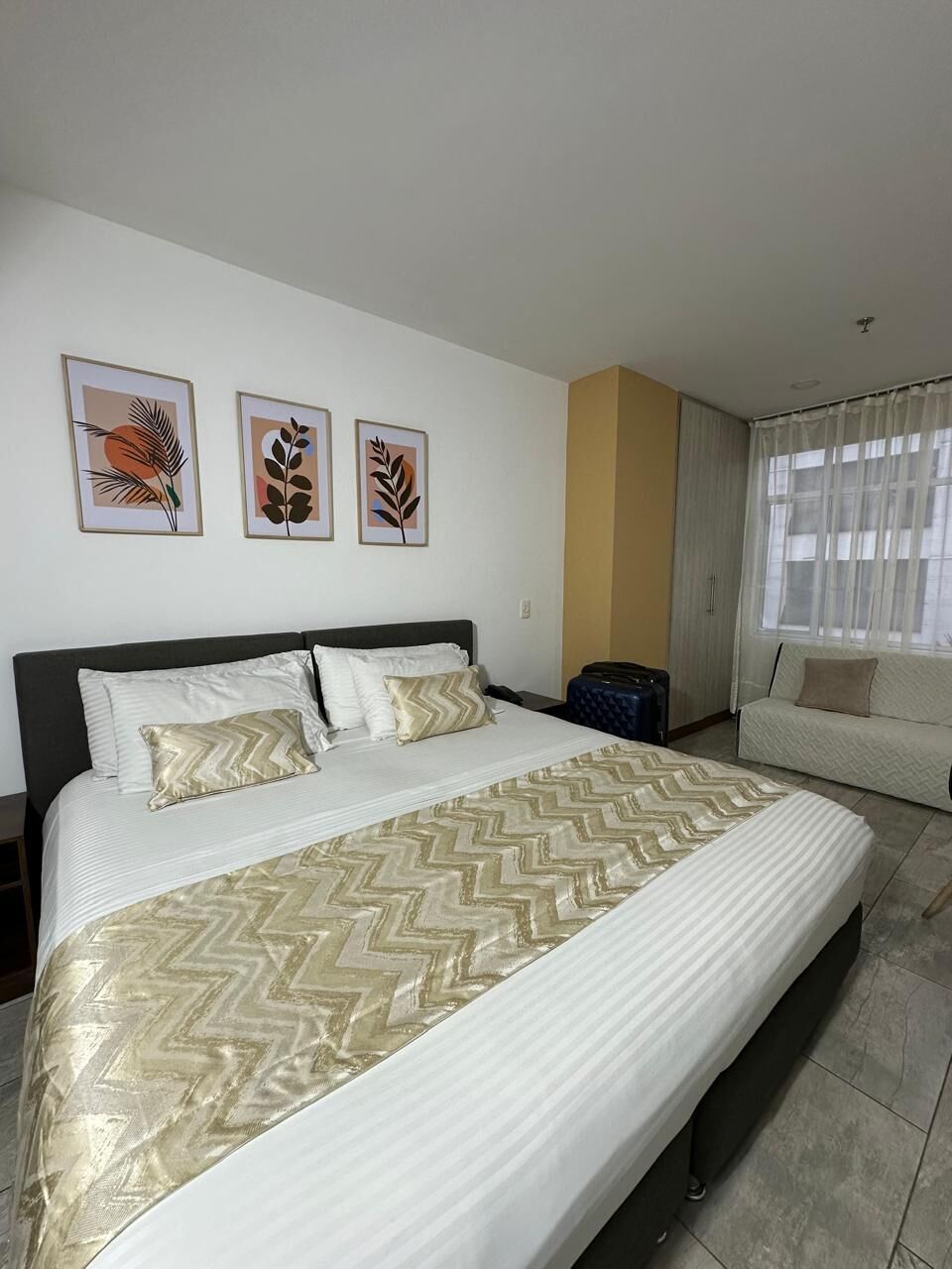 A hotel room with a large bed, a sofa, a suitcase, and three framed wall art pieces.