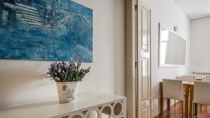 A modern room with a white console table, a potted lavender plant, a blue abstract painting, a dining area with white chairs, and a large mirror. - GuestReady - House Marquês - Ponte S. João (Porto)