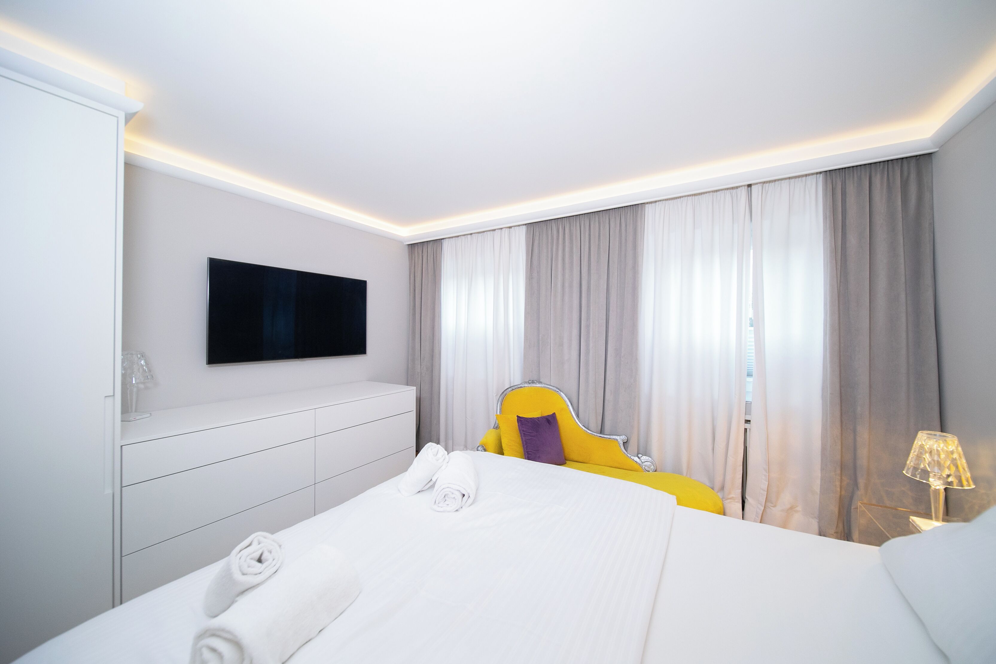 A hotel room with a bed, a TV mounted on the wall, a bedside table with a lamp, and a window with curtains.
