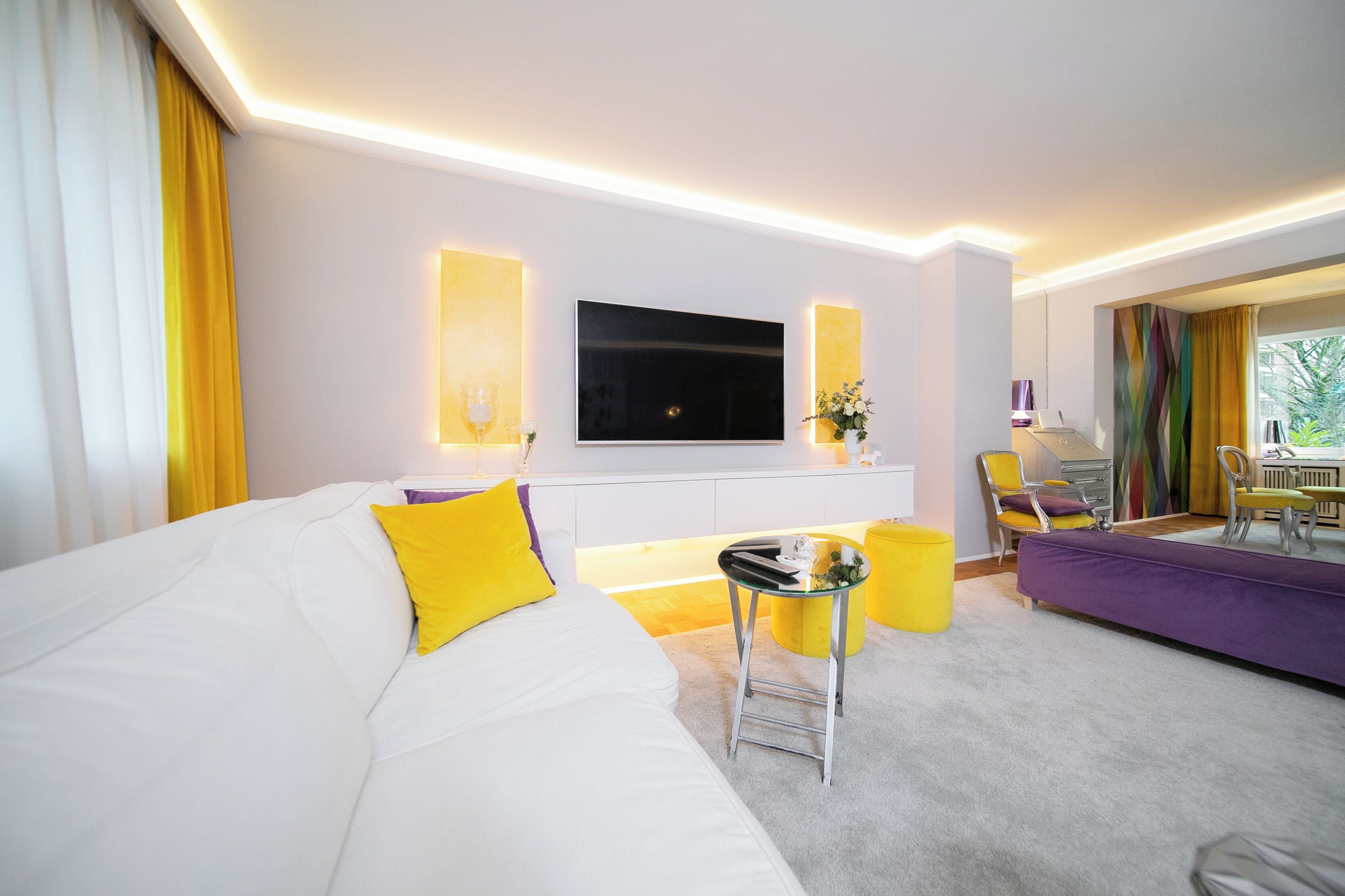 Une chambre d’hôtel moderne avec un grand canapé blanc, un téléviseur à écran plat fixé au mur, une petite table d’appoint et un canapé violet.