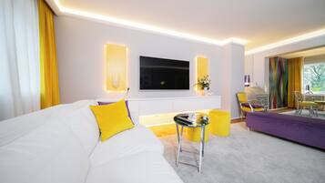 Une chambre d’hôtel moderne avec un grand canapé blanc, un téléviseur à écran plat fixé au mur, une petite table d’appoint et un canapé violet.