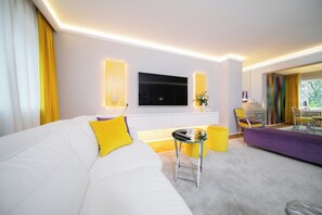 A modern hotel room with a large white sofa, a flat-screen TV mounted on the wall, a small side table, and a purple sofa. - Favorite Stays - Suite and More - Deluxe (Neuss)