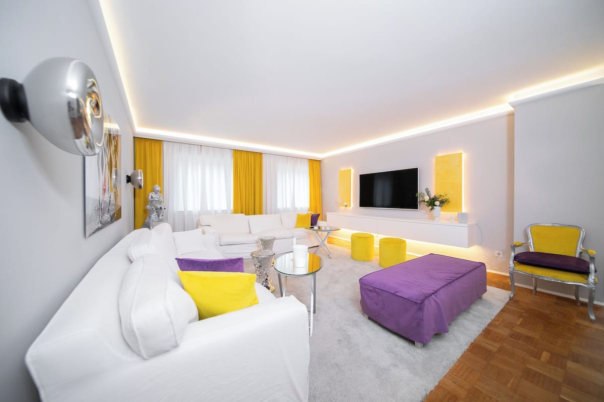 Uma sala de estar moderna com um sofá branco, um banquinho roxo e detalhes em amarelo.