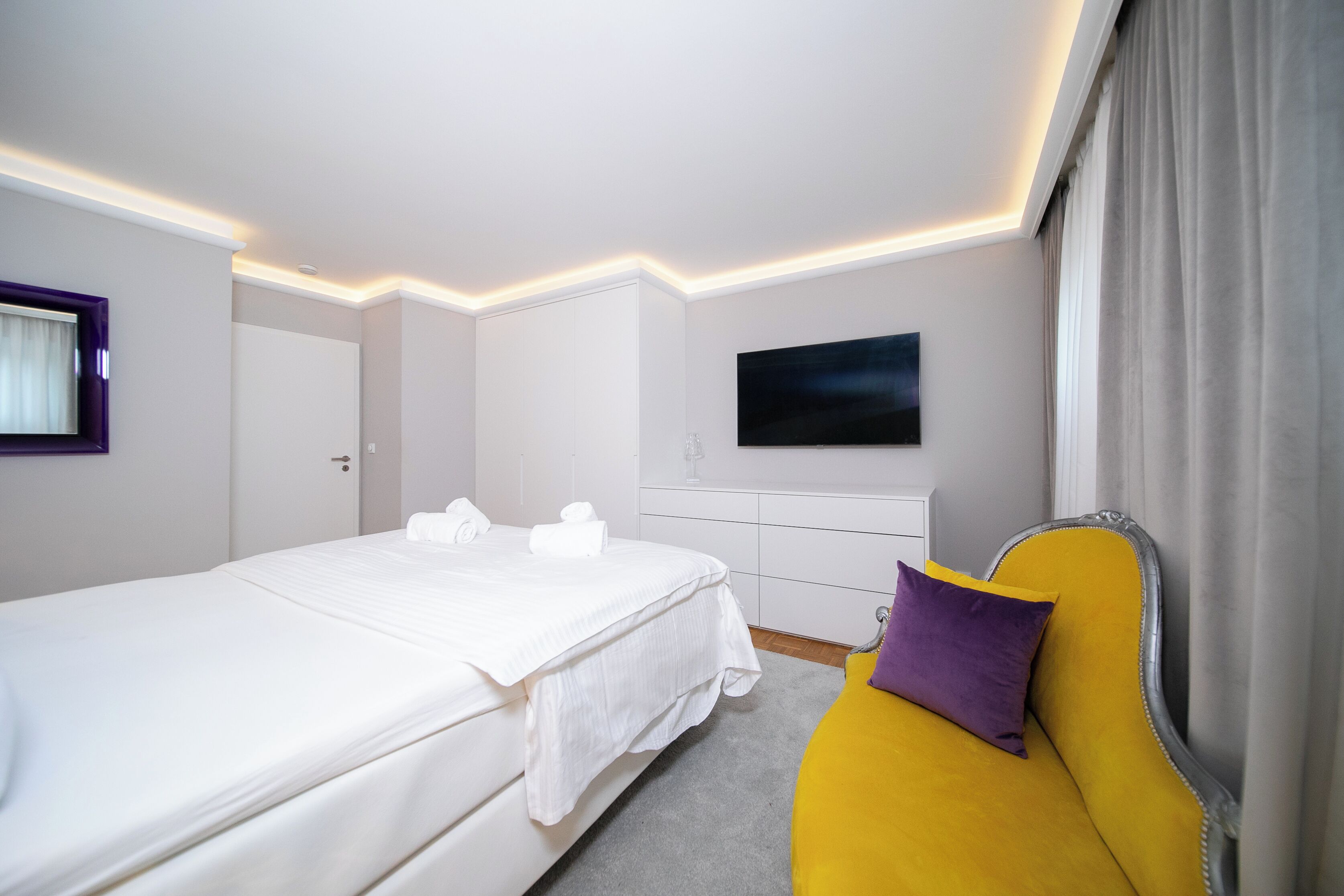A hotel room with a bed, a yellow sofa, a flat-screen TV, and a window with curtains.