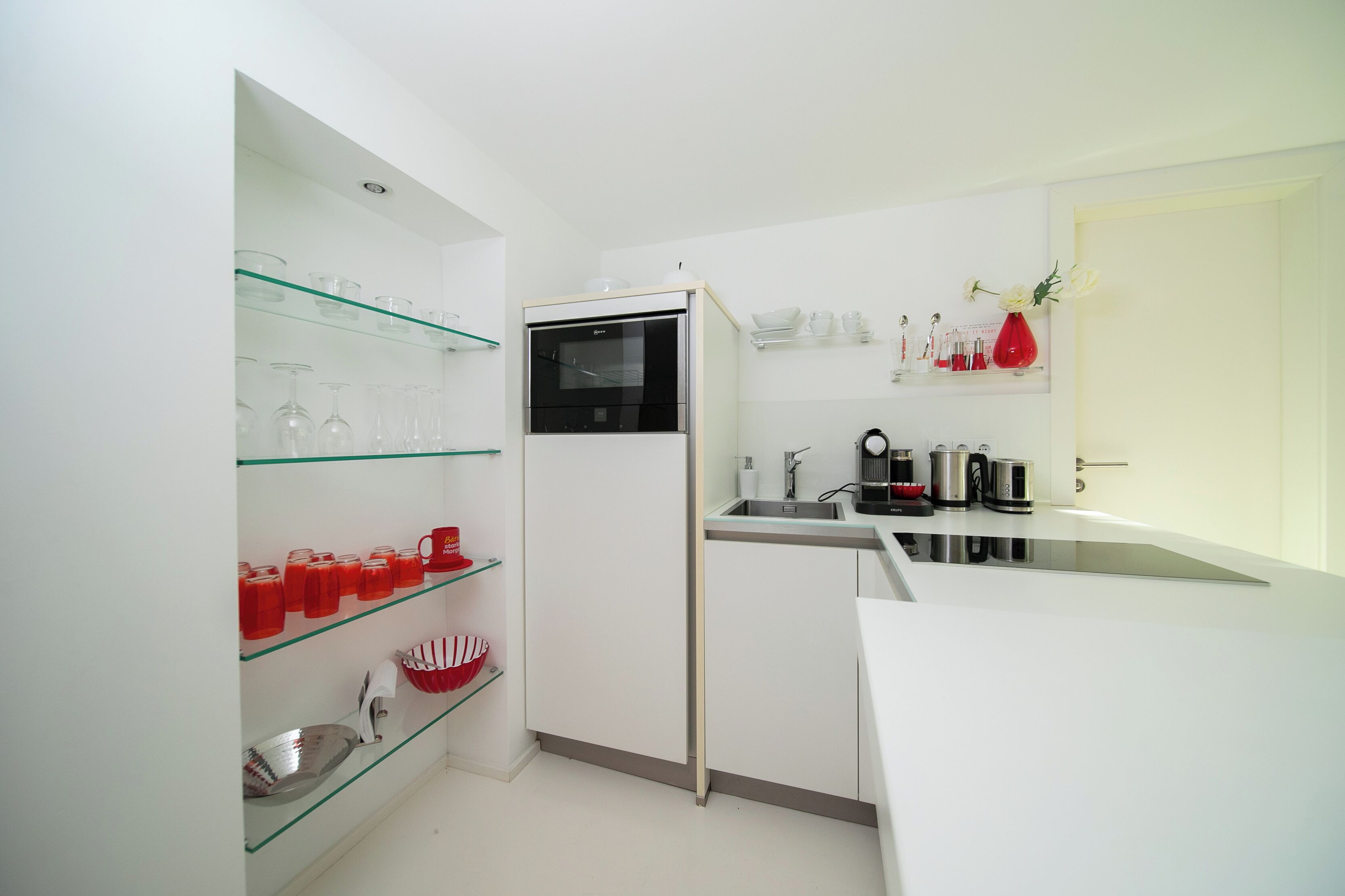 A compact kitchen with white cabinets, a built-in oven, and glass shelves holding kitchenware.