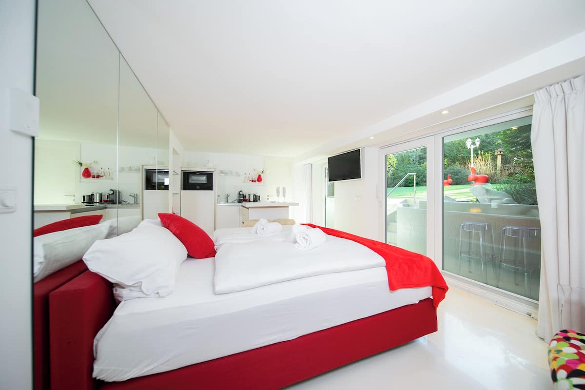 Quarto de hotel moderno com uma cama grande, televisão, varanda com área de estar e vista para o jardim.
