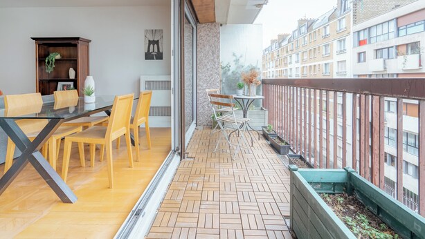 Un balcon doté d’un plancher en bois, d’une table en verre et de chaises. On peut apercevoir des bâtiments au loin.