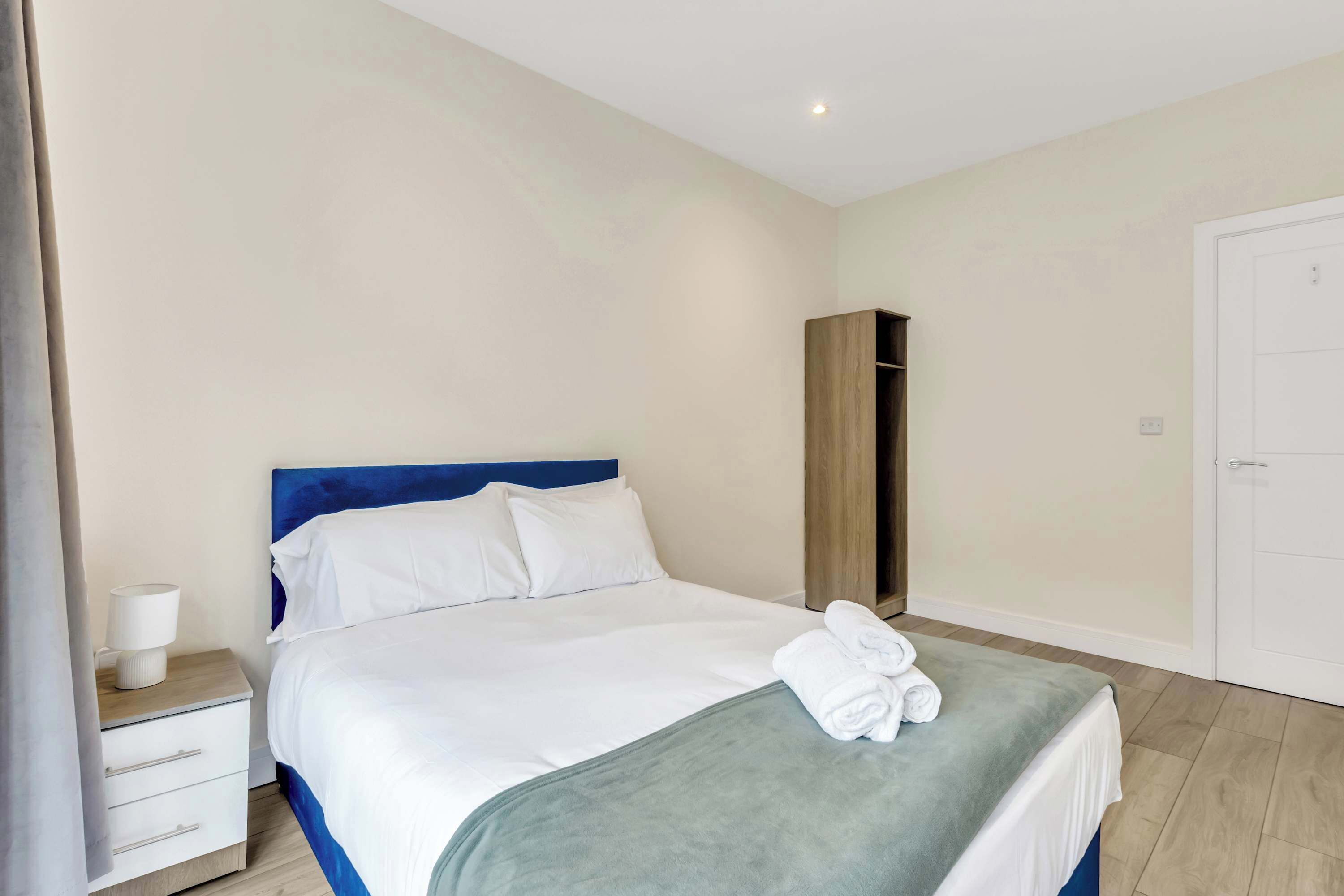 A neatly made bed with white linens and a blue headboard, a wooden wardrobe, a white bedside table with a lamp, and a white door.