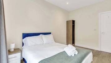 A neatly made bed with white linens and a blue headboard, a wooden wardrobe, a white bedside table with a lamp, and a white door.