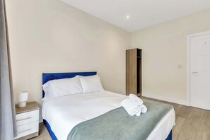 Cama bem arrumada com lençóis brancos e cabeceira azul, guarda-roupa de madeira, mesa de cabeceira branca com abajur e porta branca.