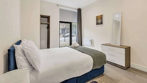 A modern bedroom with a large bed, a bedside table, a dresser, and a window with a view of the street.