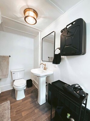 A bathroom with a white toilet, a pedestal sink, a mirror, and a black cabinet with a towel hanging on it. - Charming Mtn Stay | Views, Hot Tub, Prime Location (West Kill)